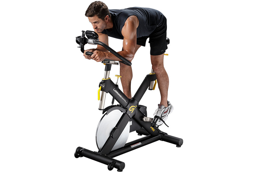 Man in black sleeveless shirt and shorts riding black and white indoor stationary bike with yellow accents on white backgroun