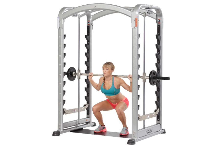 Woman in teal sports bra and red shorts performing squat on silver and black Hoist Smith Machine with barbell loaded with wei