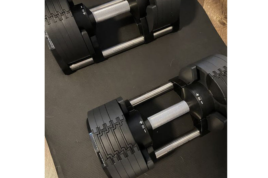 Pair of matte black adjustable dumbbells with metal handles and numbered weight plates on gray mat, flat lay view