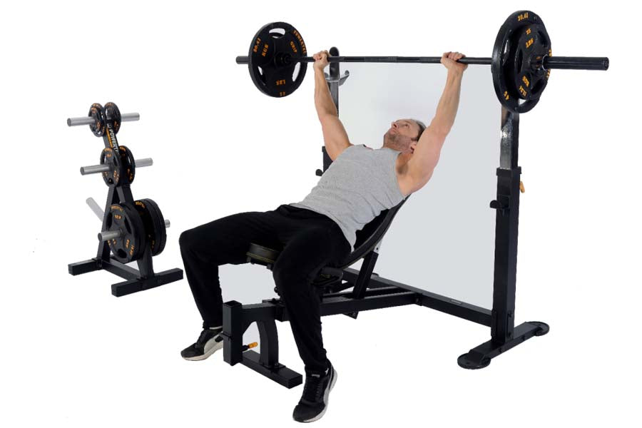 Man performing barbell bench press on black Powertec Olympic weight bench with gold-accented weight plates, dumbbell rack bes