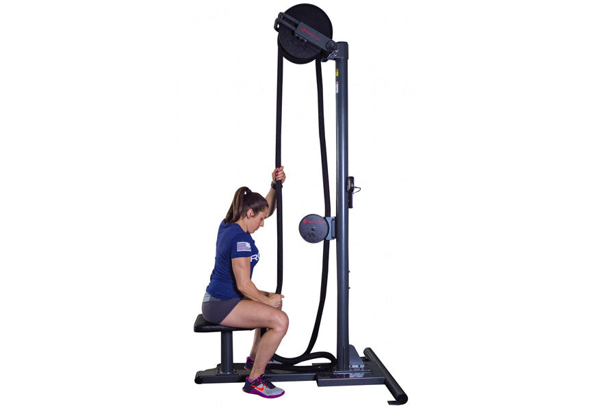 Woman using black upright rope trainer machine with dual pulleys, seated on bench, on white background