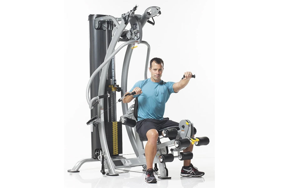Man in blue shirt using silver and black multi-station home gym machine with adjustable arms and leg press on white backgroun