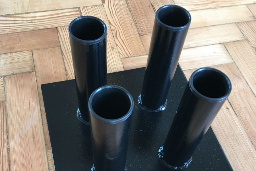 Black metal vertical barbell holder with five cylindrical tubes mounted on square base, displayed on wooden floor
