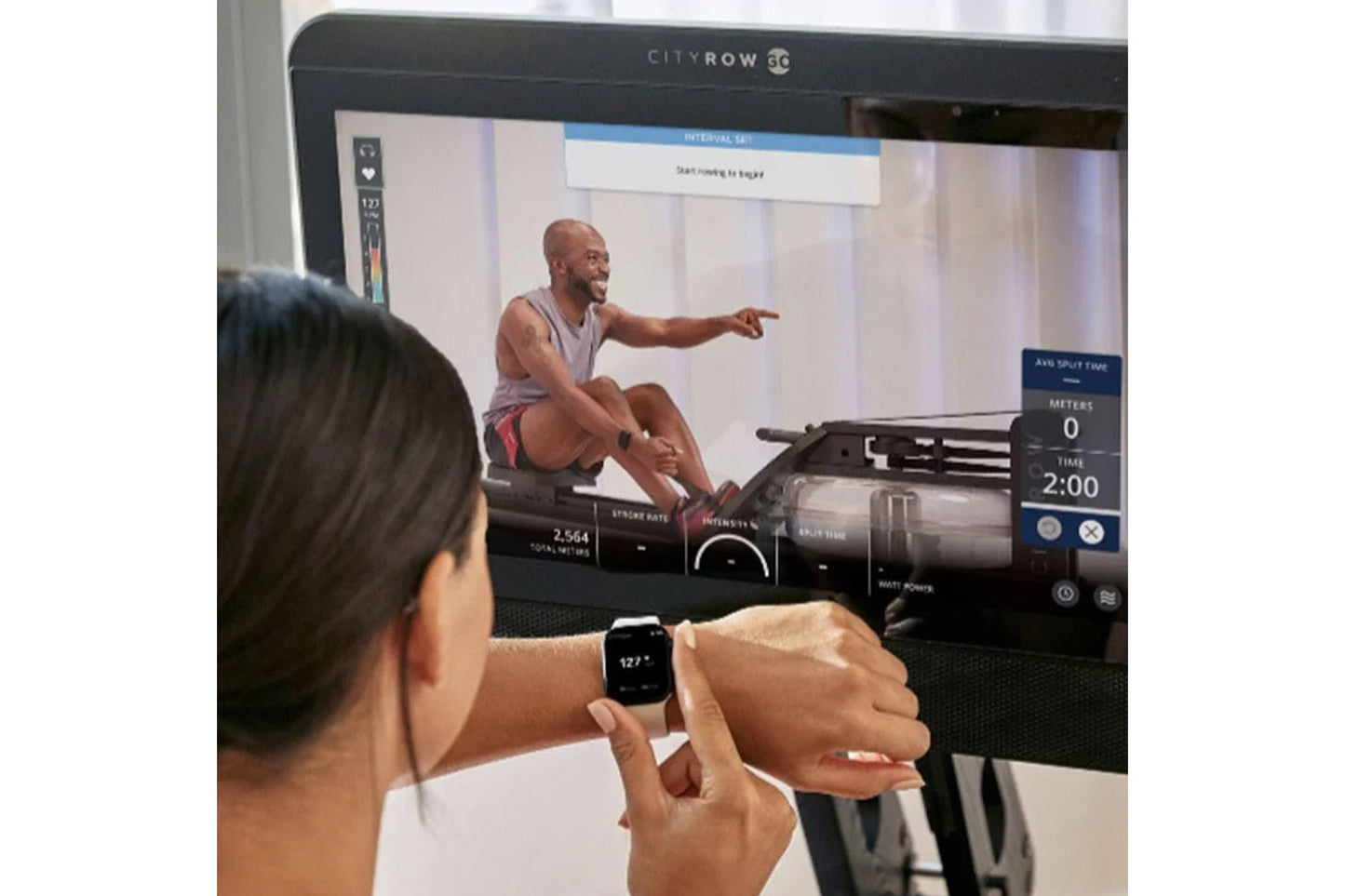 Person wearing smartwatch viewing CityRow GC rowing machine display screen showing instructor-led workout with performance me