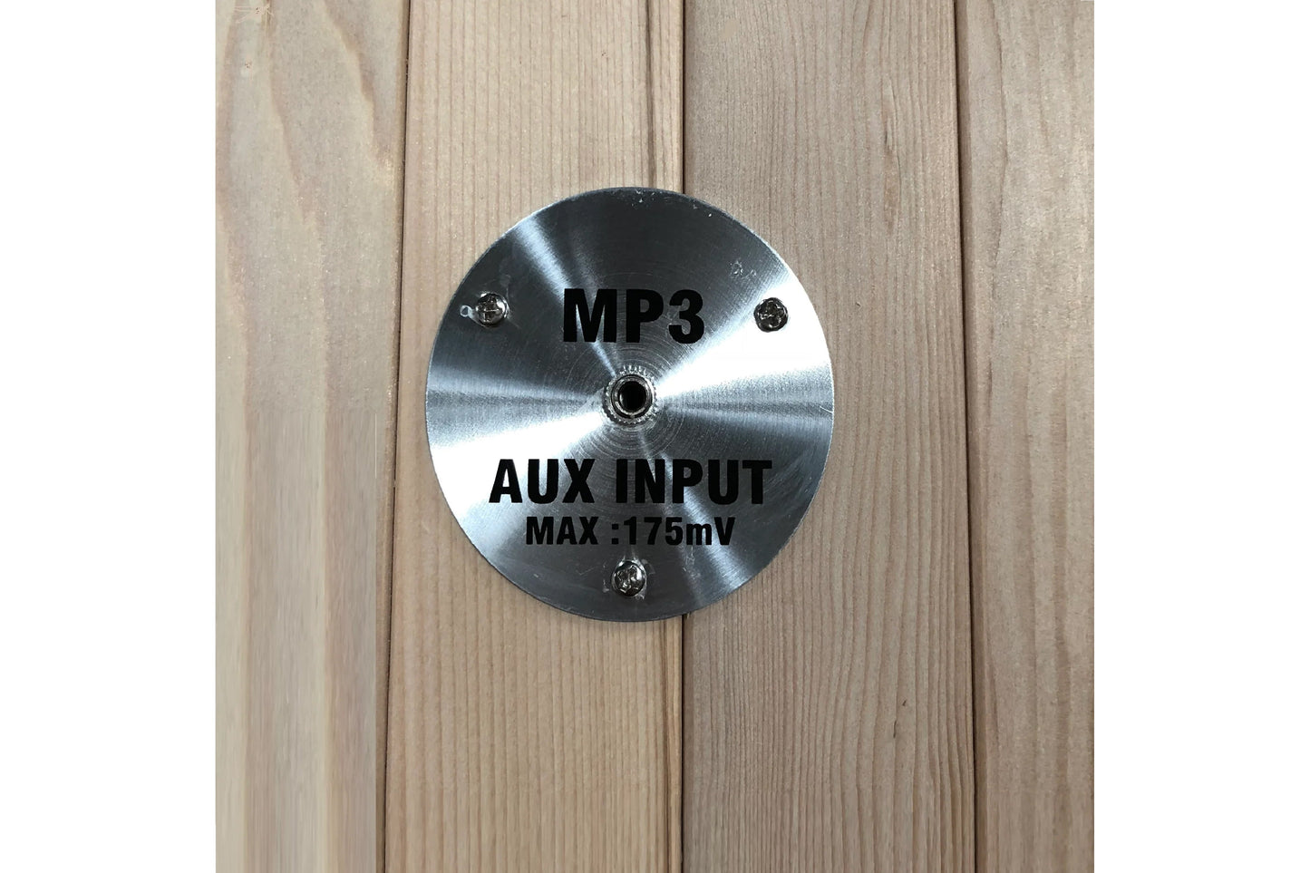 Brushed silver circular control plate with black text reading "MP3" and "AUX INPUT MAX :175mV" mounted on light wood sauna in