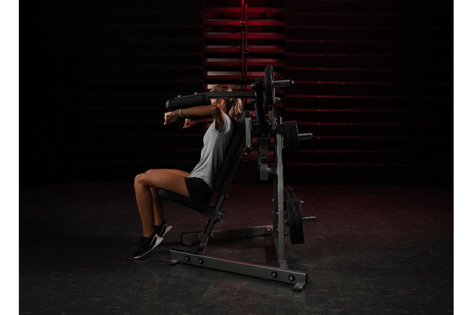 Woman using Hammer Strength plate-loaded lateral raise machine with gray tank top and black shorts in gym setting