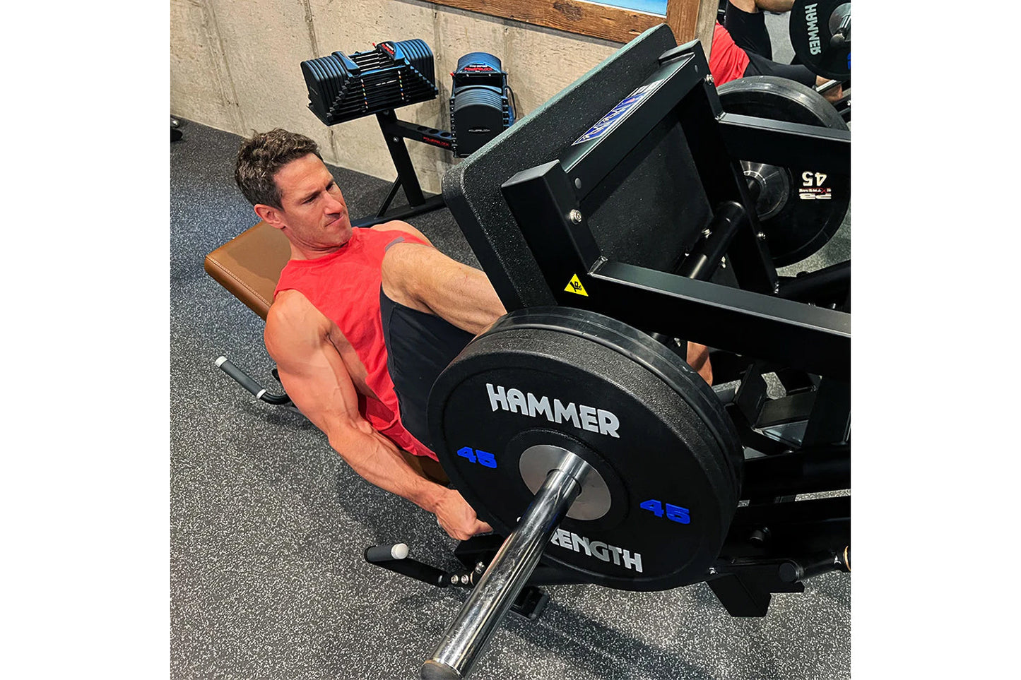 Man in red sleeveless shirt using black Hammer Strength plate-loaded leg press machine in gym setting
