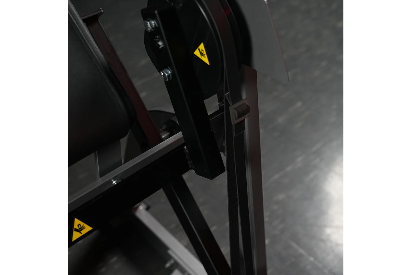 Black plate-loaded seated biceps machine with yellow caution labels and metal frame, detail view of mechanical arm and handle