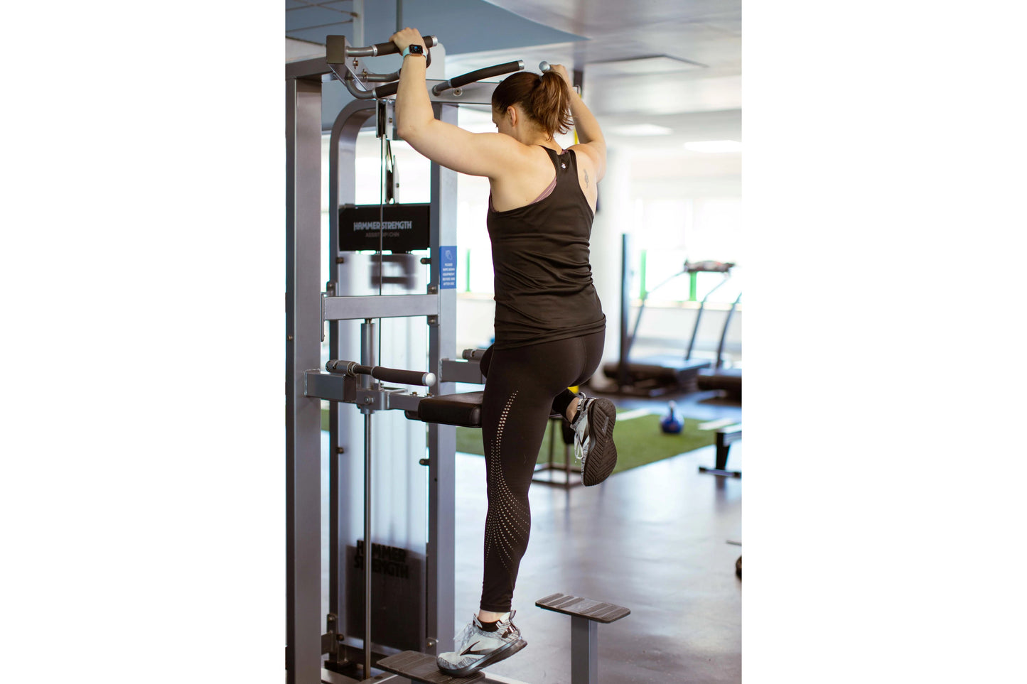 Woman performing assisted dip exercise on Hammer Strength Select machine in gym setting, wearing black sports bra and legging