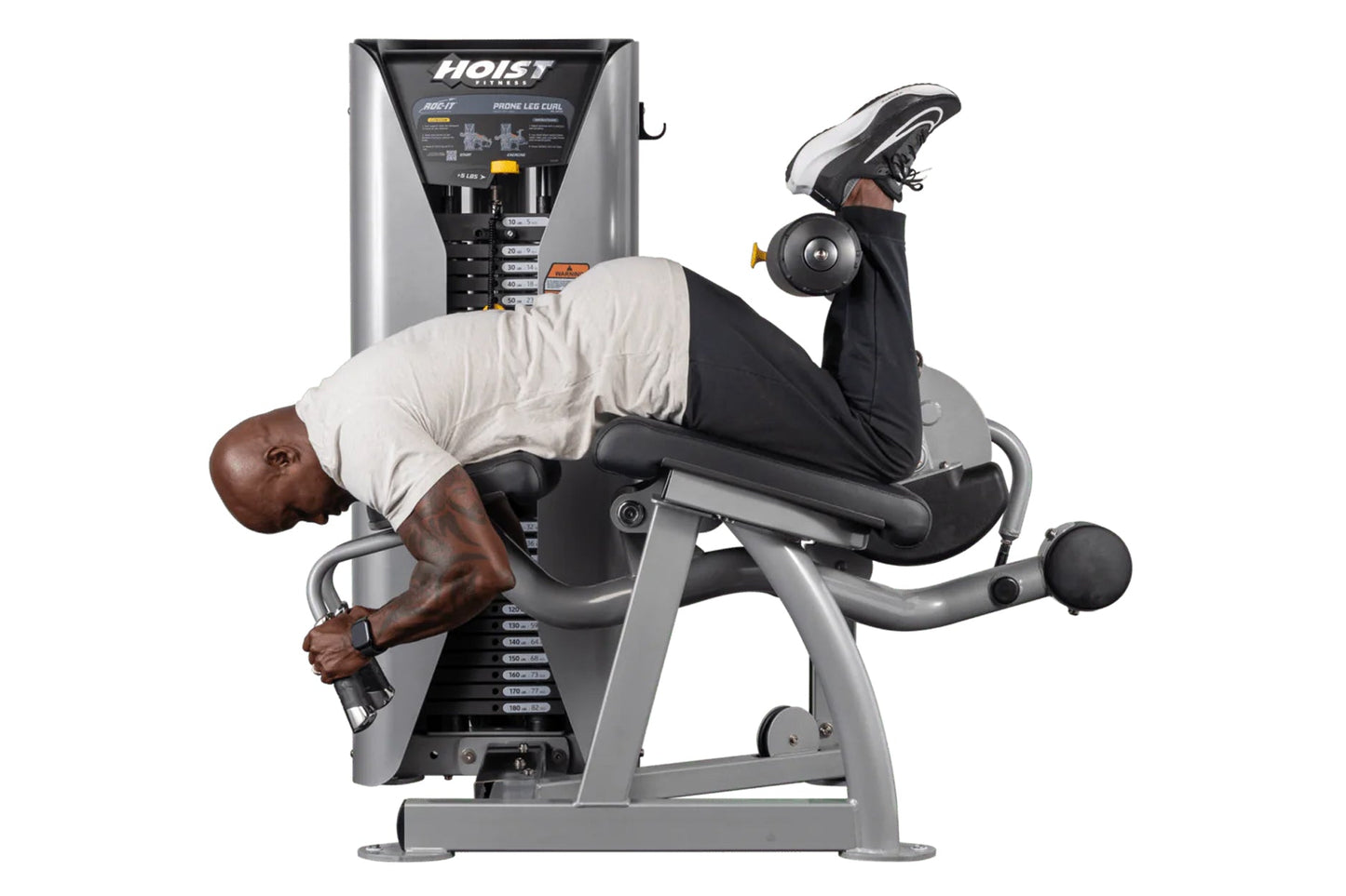 Man using Hoist ROC-IT prone leg curl machine with silver frame and black padding, holding dumbbell
