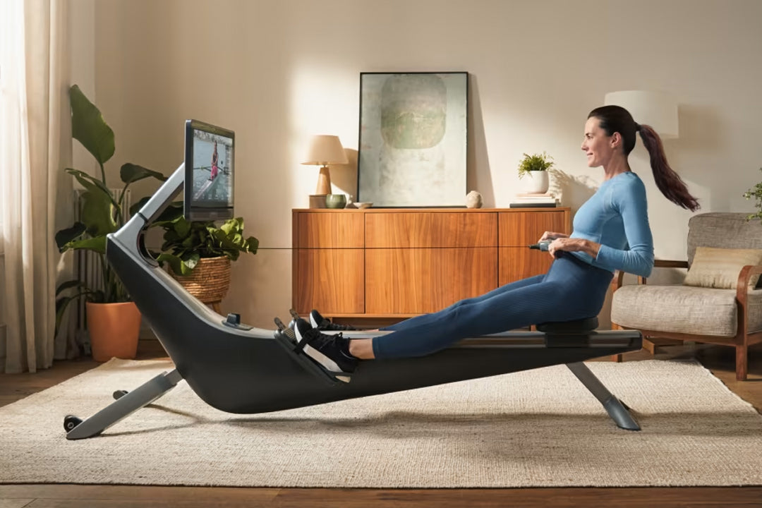 Woman in blue long-sleeve shirt and navy leggings using black Hydrow Pro rowing machine in home gym setting with wood dresser