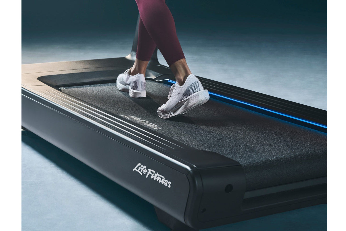 Person wearing burgundy cropped leggings and white mesh running shoes jogging on a black Life Fitness treadmill with blue acc