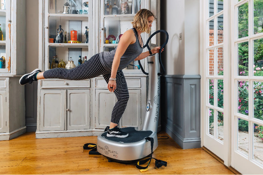 Woman in gray tank top and striped black leggings using silver Power Plate My5 vibration trainer in home gym setting