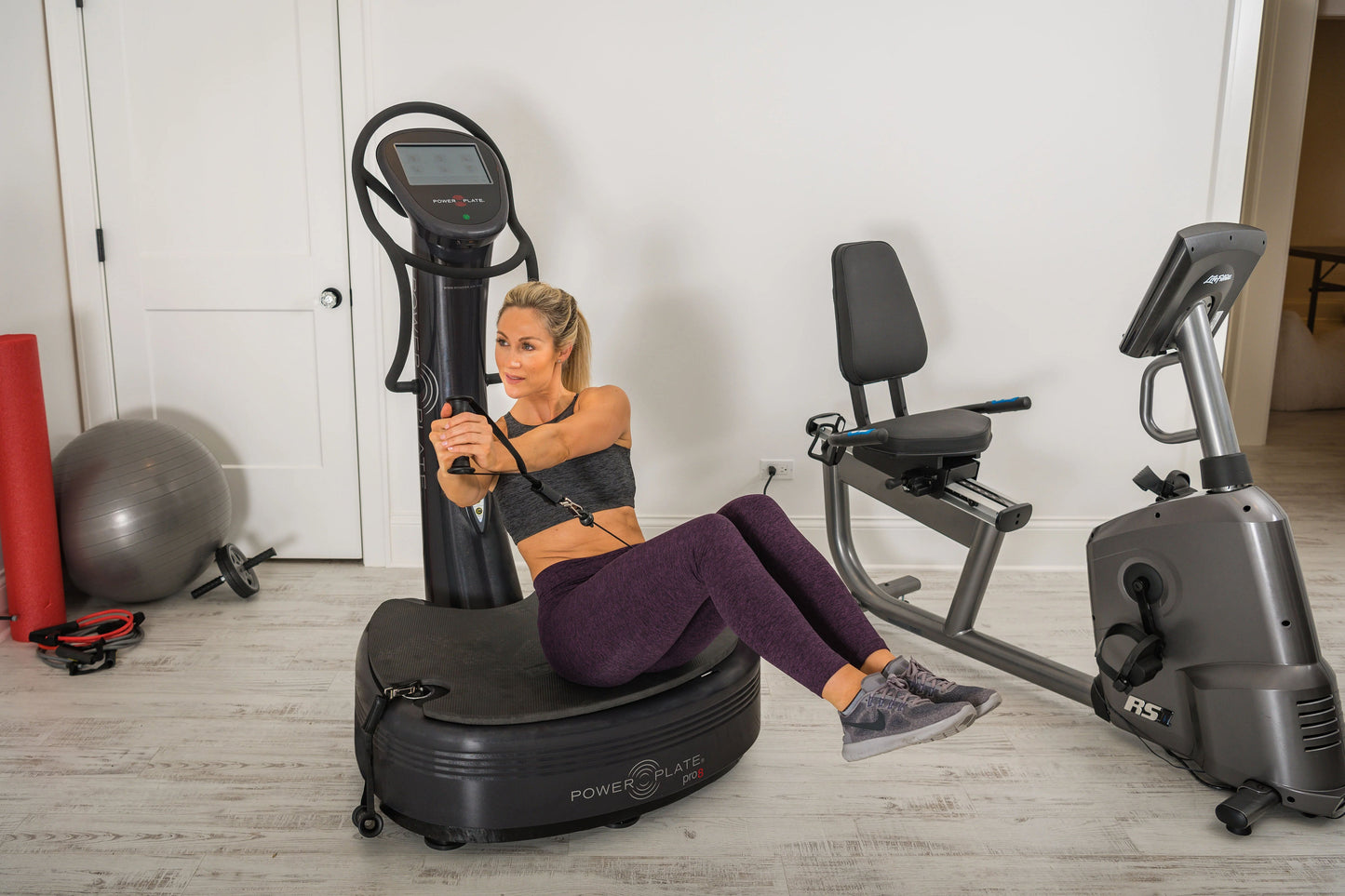 Woman using black Power Plate Pro8 vibration plate trainer in gym setting with exercise equipment