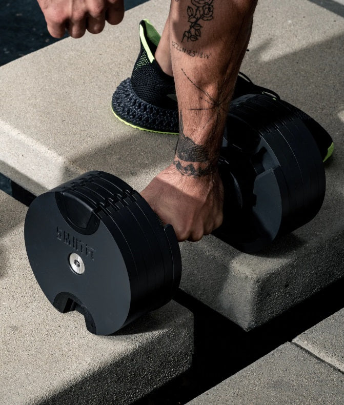 Person in black and neon green athletic shoes lifting black adjustable dumbbell on concrete steps