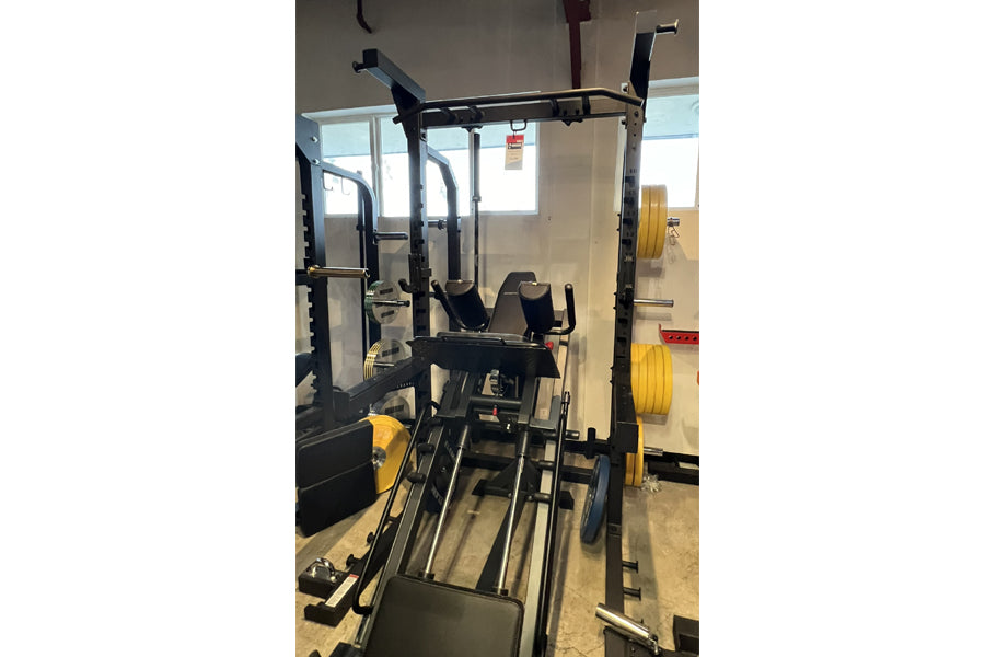 Black metal squat rack with yellow accents, vertical uprights, safety bars, and attached plate storage in gym setting