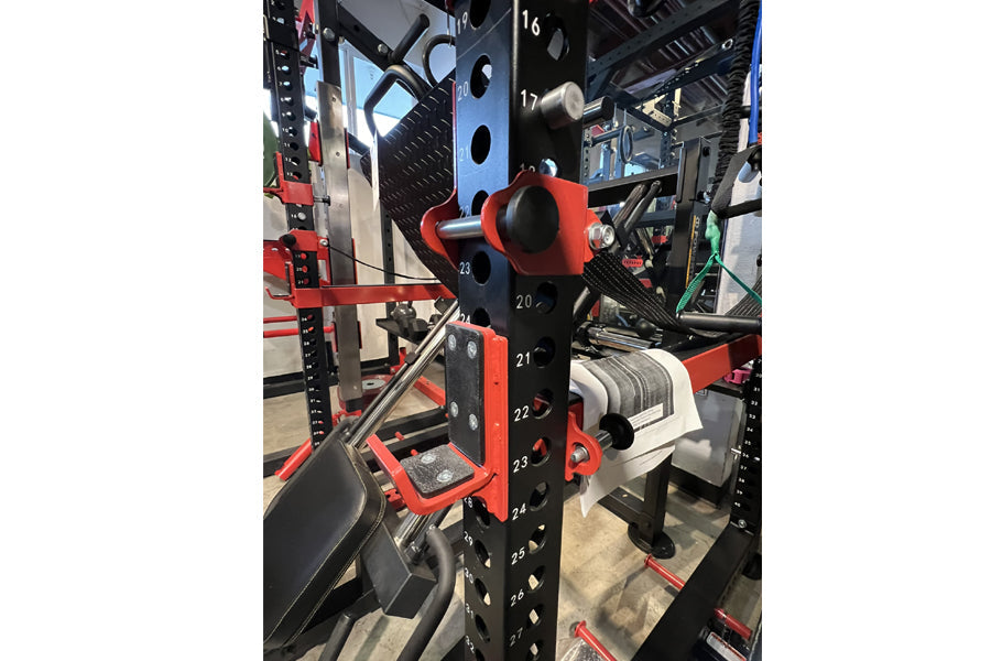Red and black weightlifting cage with numbered attachment points, featuring pull-up bar and plate storage, in gym setting