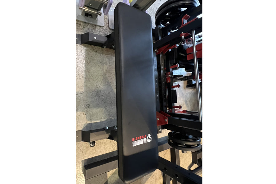Black flat weight bench with red Warrior branding and locking wheels, shown in warehouse setting next to 3D printer