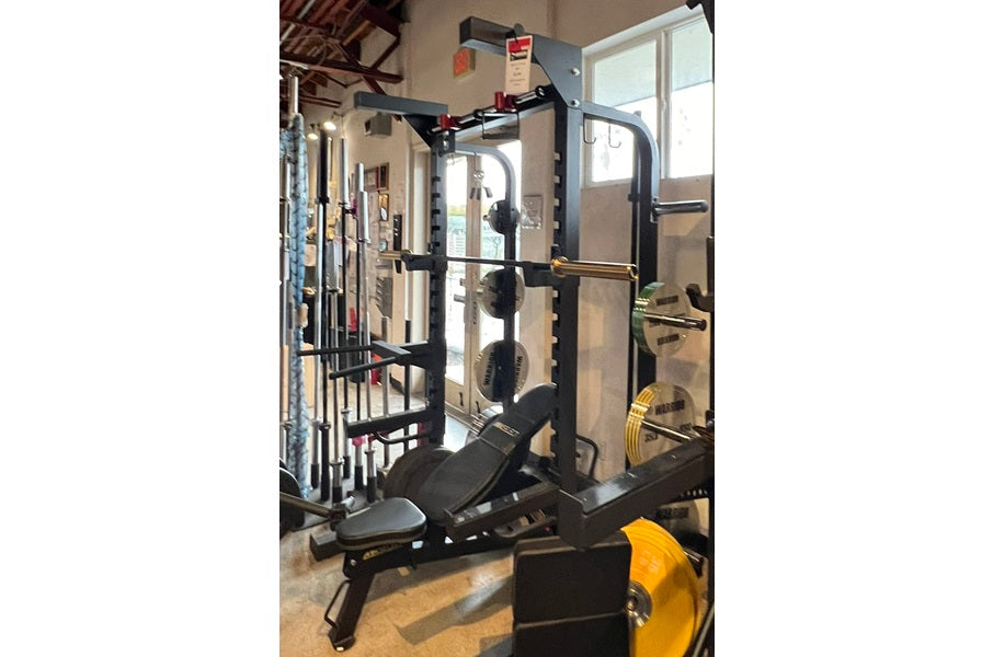 Black and gray squat rack with pull-up bars and weight storage, displayed in a gym setting with yellow weight plates visible