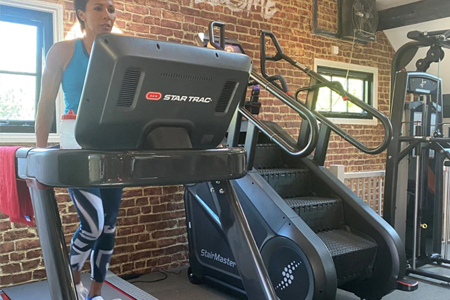 Person wearing blue tank top and patterned leggings using Star Trac treadmill in brick gym, StairMaster equipment visible nea