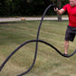 Black twisted battle ropes laid out on grass, held by man in red shirt during outdoor workout