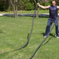 Man holding black battle ropes outdoors on grass, arms raised in exercise position, wearing navy sleeveless shirt