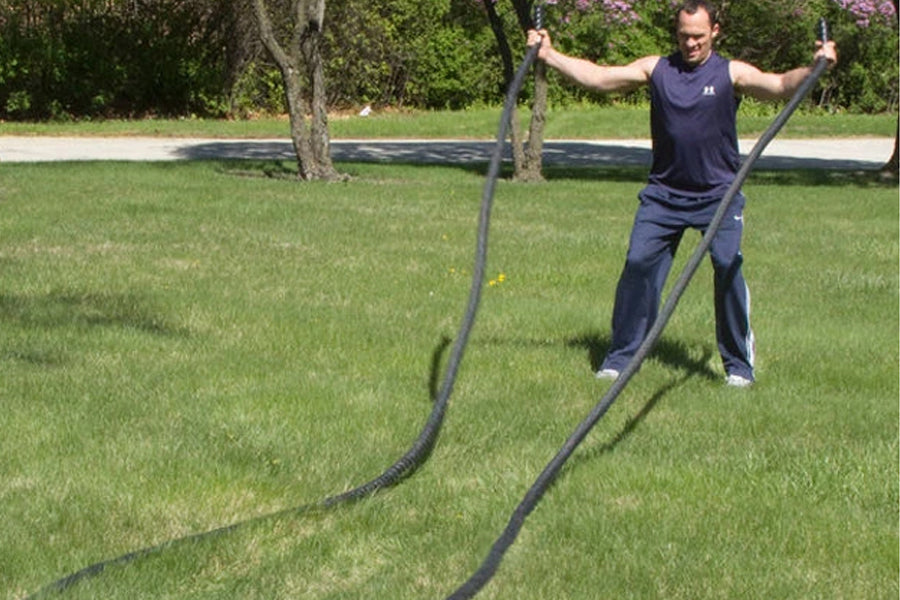 Man holding black battle ropes outdoors on grass, arms raised in exercise position, wearing navy sleeveless shirt