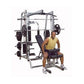Man wearing yellow tank top and purple shorts seated on Body-Solid Series 7 Smith Machine with silver frame and black weight 