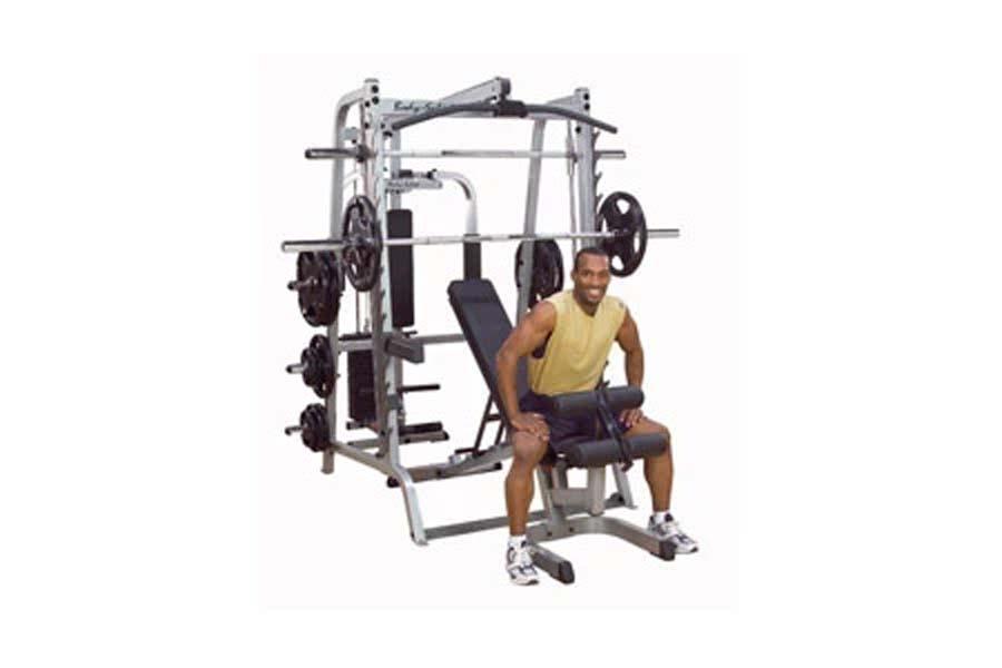 Man wearing yellow tank top and purple shorts seated on Body-Solid Series 7 Smith Machine with silver frame and black weight 