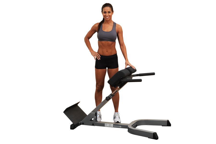 Woman wearing gray sports bra and black shorts posing with Body-Solid 45-degree hyperextension machine on white background