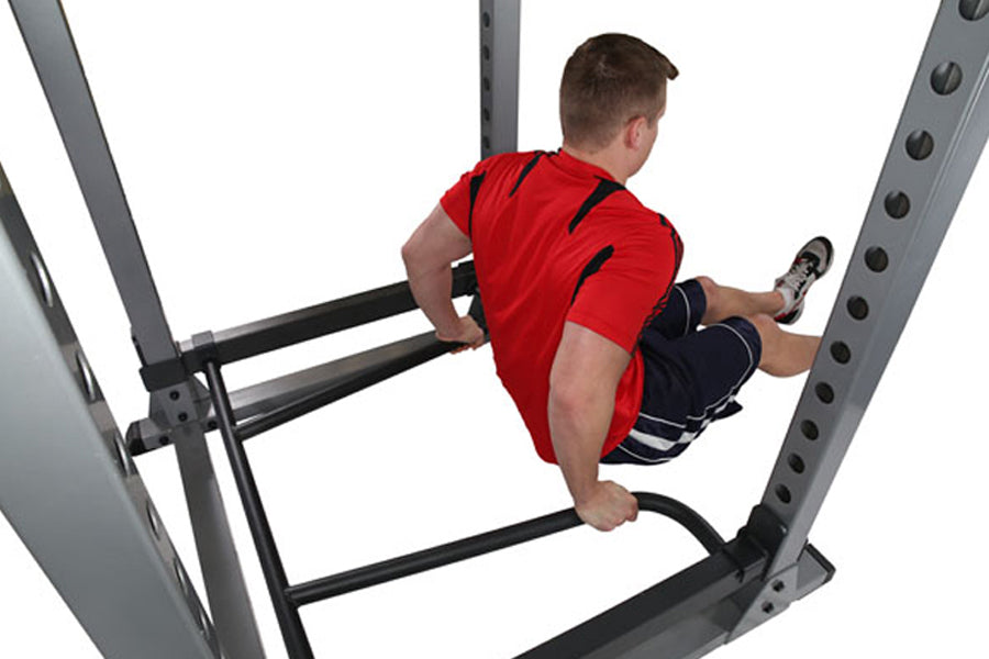 Man wearing red shirt and black shorts using dip attachment on gray metal weight rack against white background