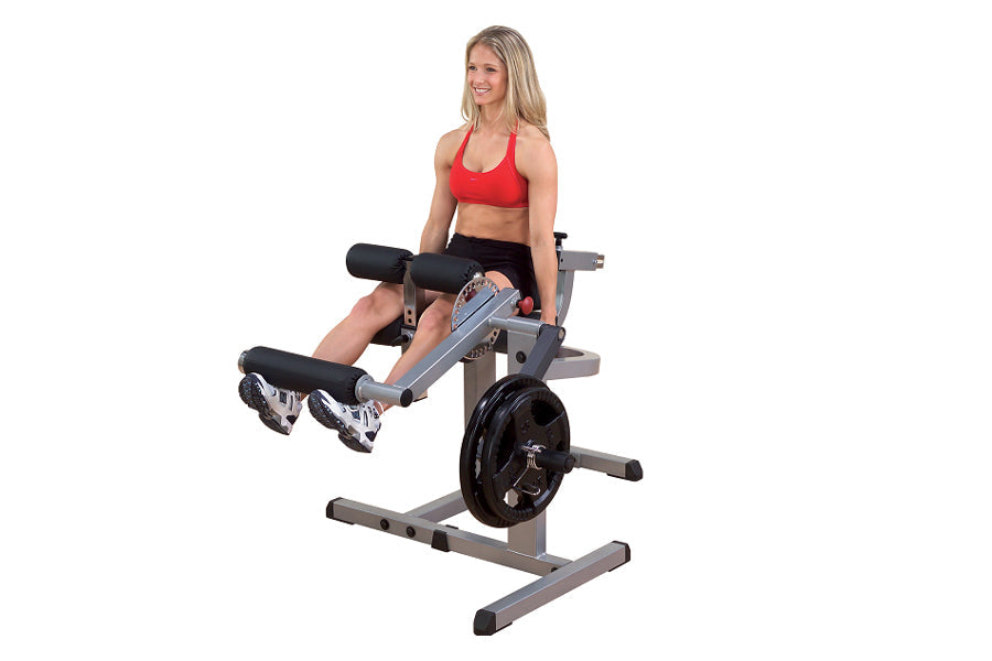 Woman in red sports bra and black shorts using gray and black leg extension machine with weighted plate, on white background