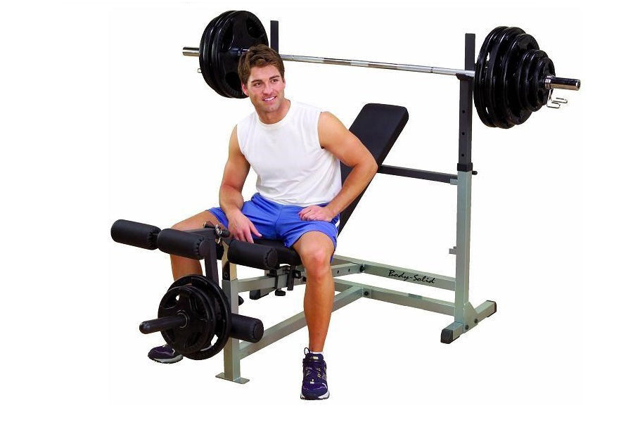 Male model wearing white sleeveless shirt and blue shorts seated on gray and black weight bench with barbell and leg extensio