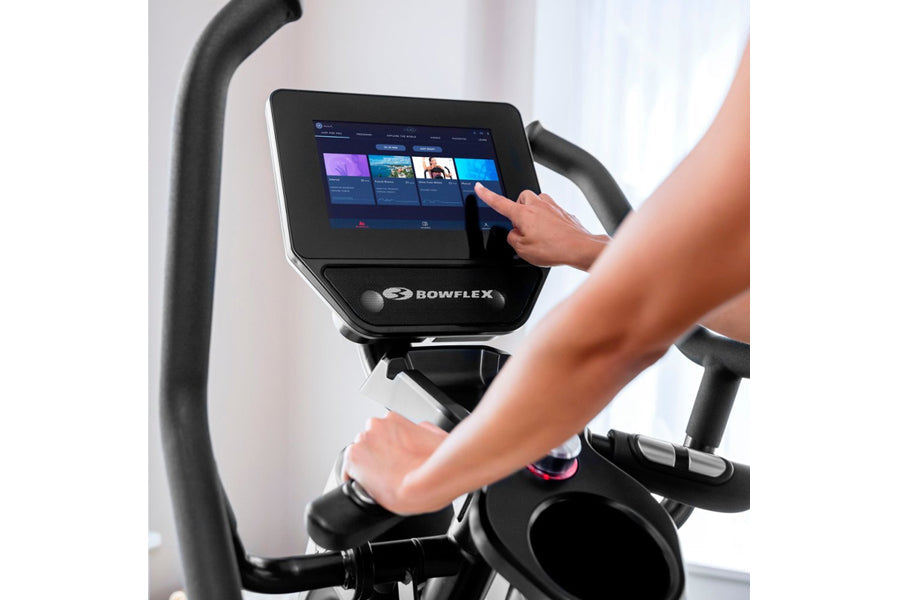 User interacting with Bowflex Max Trainer M9 elliptical machine with touchscreen display showing fitness app interface
