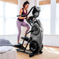 Woman in purple sports bra and leggings using black Bowflex Max Trainer M9 elliptical machine in bright home gym with wooden 