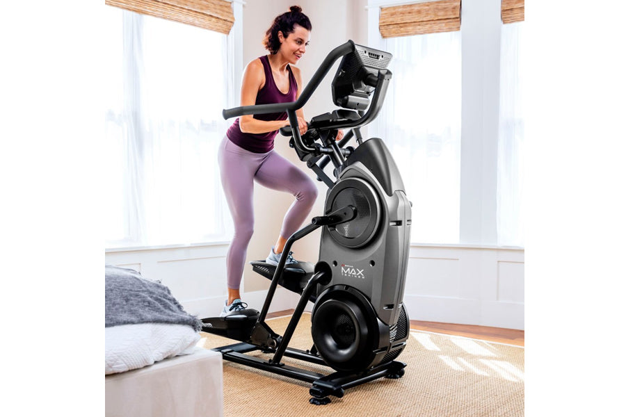 Woman in purple sports bra and leggings using black Bowflex Max Trainer M9 elliptical machine in bright home gym with wooden 