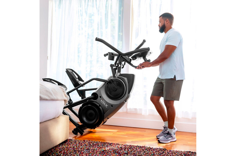 Man in light blue t-shirt and gray shorts using black Bowflex Max Trainer M9 elliptical machine in bright home gym with windo