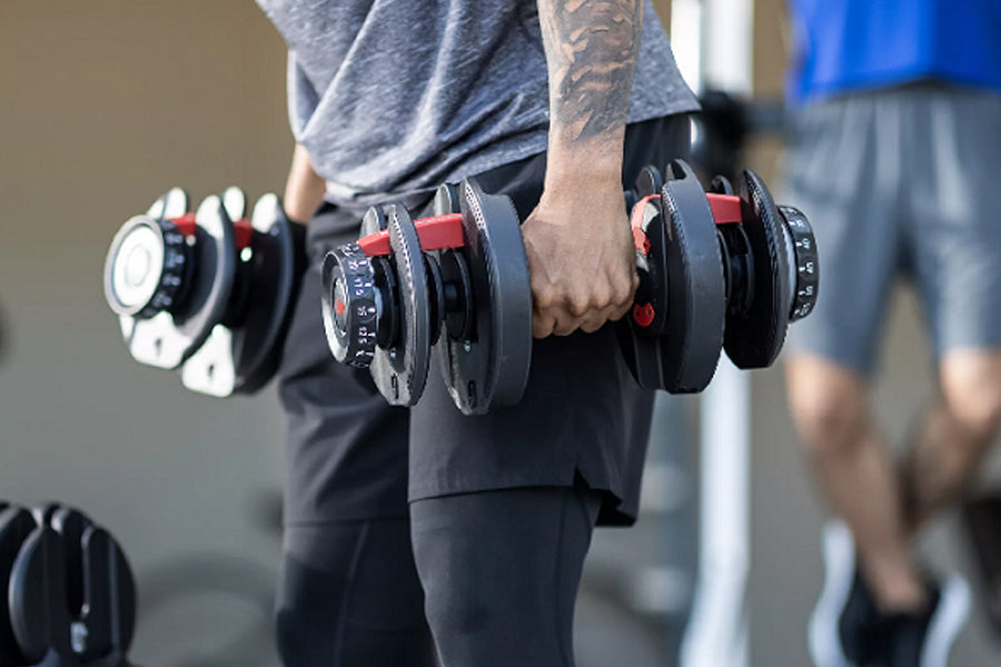 Person holding pair of black and red adjustable dumbbells with numbered weight dial, gym setting