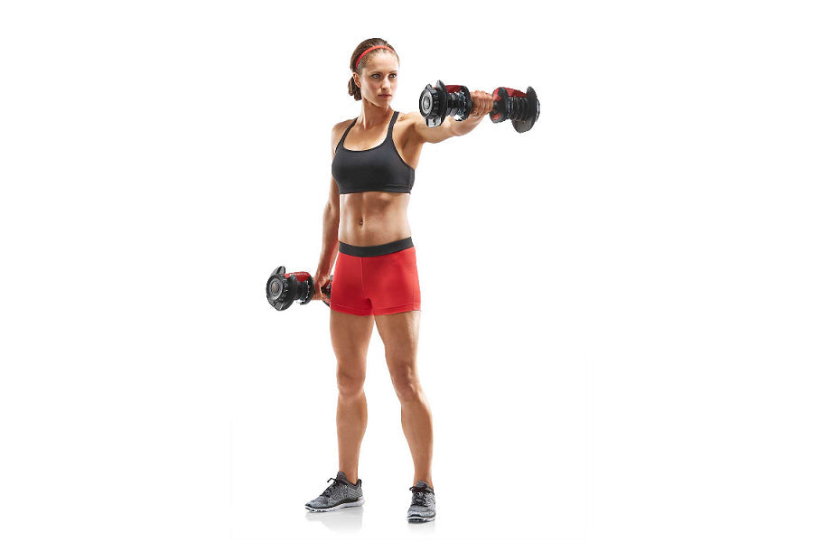 Athletic woman wearing black sports bra and red shorts holding red and black adjustable dumbbells in shoulder press position 