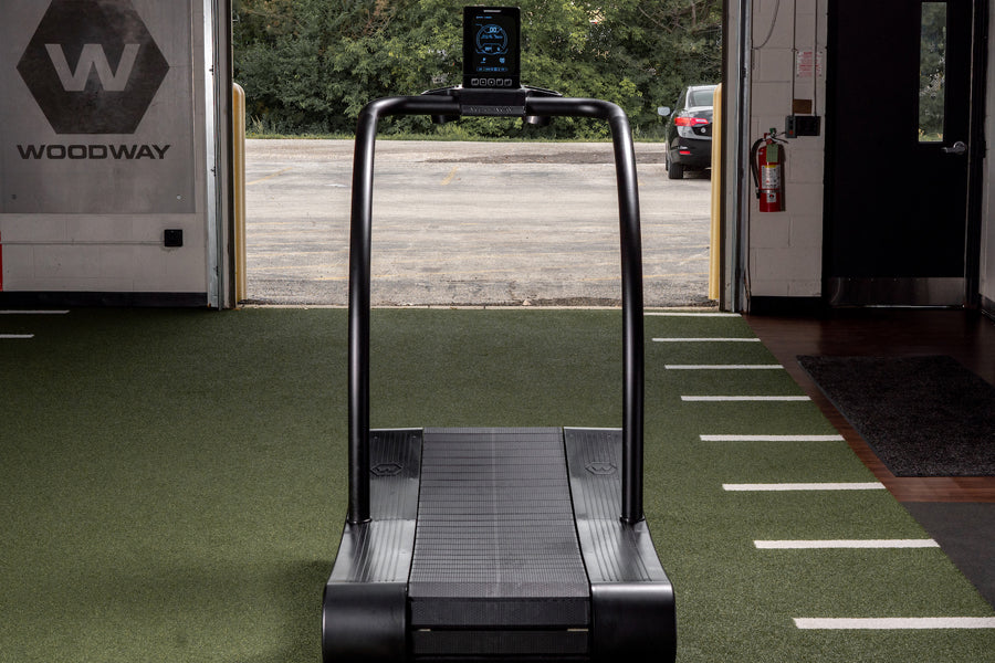 Black curved treadmill with digital display mounted on arch, shown on synthetic turf in fitness showroom