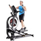 Man exercising on black and gray California Fitness elliptical machine with red wheel, wearing blue tank top and black shorts