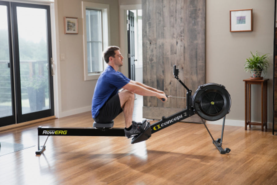 Man in blue shirt using black Concept2 RowErg rowing machine in modern home gym with wood flooring