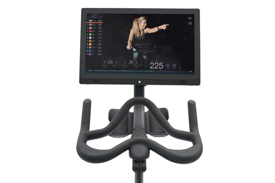 Black indoor exercise bike with digital console displaying workout metrics and video instruction from trainer on white backgr