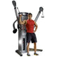 Man in red tank top and black shorts demonstrating dual cable cross machine with adjustable arms and weight stack on white ba