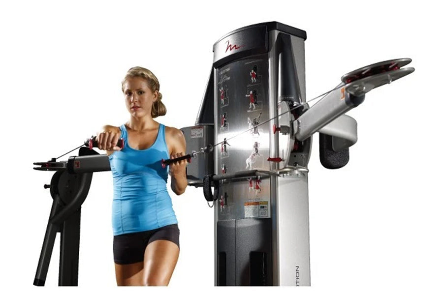 Woman in blue tank top and black shorts using dual cable crossover exercise machine with adjustable arms on white background