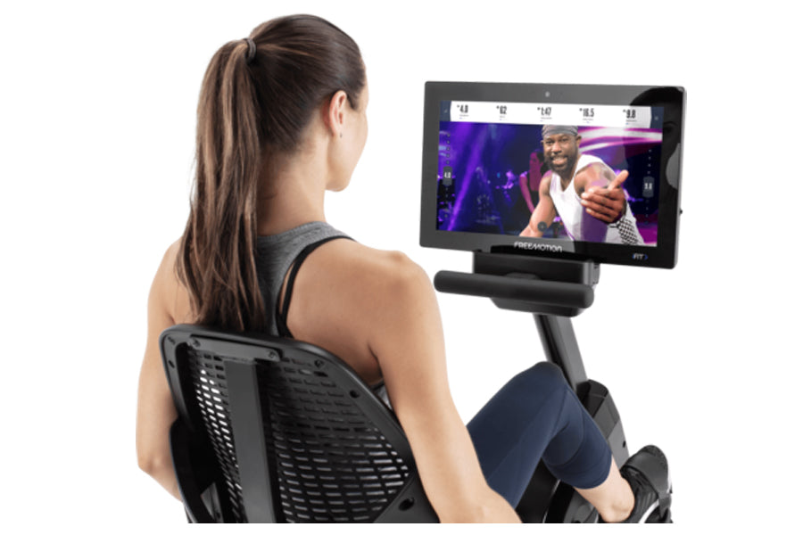 Woman exercising on black recumbent bike with digital display screen showing fitness video on white background