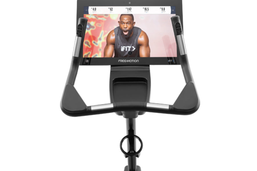 Black upright exercise bike with tablet display screen showing fitness content, gray handles, and pedal base on white backgro