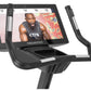 Black Freemotion u22.9 upright exercise bike with tablet display showing fitness instructor, adjustable handlebars, and pedal