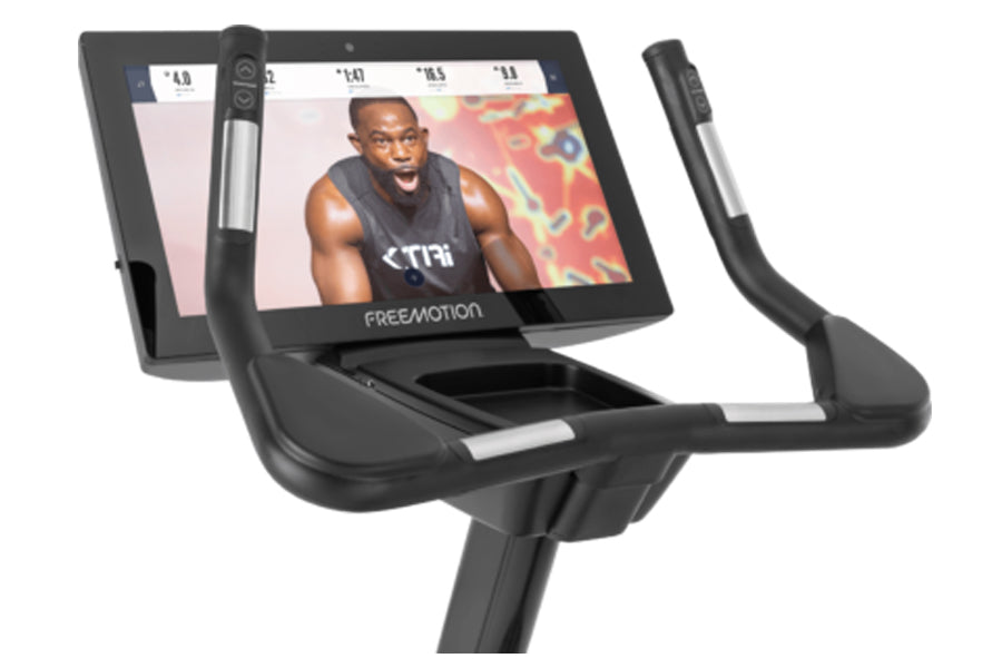 Black Freemotion u22.9 upright exercise bike with tablet display showing fitness instructor, adjustable handlebars, and pedal