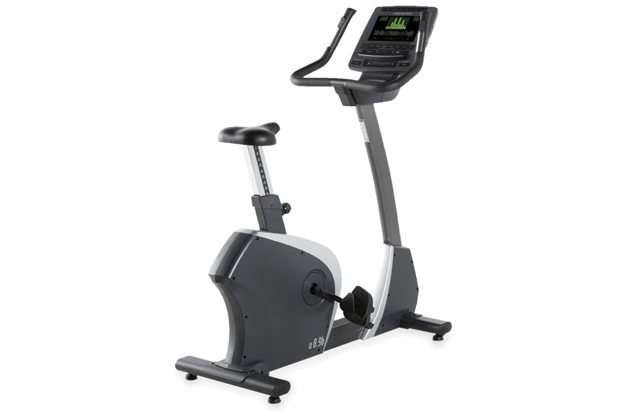 Black and white upright exercise bike with digital display console showing green metrics, foot pedals, and adjustable seat on